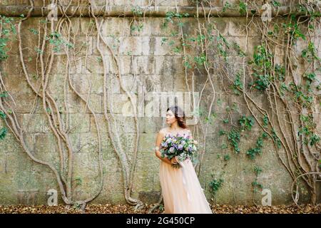 Bella sposa in un abito da sposa beige con una bella bouquet di fiori sullo sfondo di un muro di pietra intrecciato con edera Foto Stock