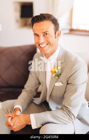 Lo sposo in un abito grigio con un arco bianco cravatta e un boutonnière con fiori arancioni siede in un camera d'albergo e sorrisi Foto Stock