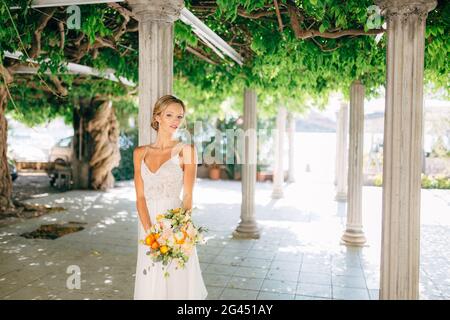 La sposa si trova tra colonne bianche intrecciate con viti verdi e tiene un bouquet di nozze con coppe d'arancia Foto Stock