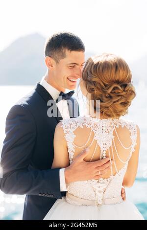 La sposa e lo sposo stanno in piedi, abbracciando e sorridendo sul molo vicino al mare, da vicino Foto Stock