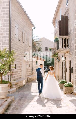 La sposa e lo sposo camminano lungo un'accogliente strada stretta della città vecchia di Perast tenendo le mani, vista posteriore Foto Stock