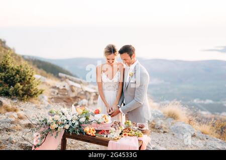 Lo sposo e lo sposo stanno tagliando una torta durante un Tavolo a buffet dopo la cerimonia nuziale sul Monte Lovcen e. sorridente Foto Stock