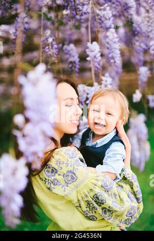 Carino bambino dai capelli rossi che ride tra le braccia della madre sotto un albero di wisteria Foto Stock