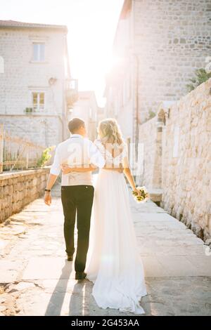 La sposa e lo sposo si abbracciano lungo le strade di Perast accanto a case di mattoni bianchi, vista posteriore Foto Stock