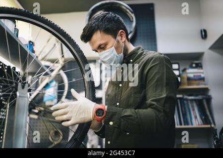 Il riparatore di biciclette lavora in officina di riparazione e assistenza biciclette durante la quarantena del coronavirus indossando visiera e guanti Foto Stock