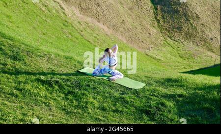 Donna yogi praticare yoga in natura, verdi colline in estate su un tappetino yoga. Una ragazza caucasica dai capelli castani in un'asana in abbigliamento sportivo, leggings e una T-shirt con capelli intrecciati in una coda di pony. Foto Stock