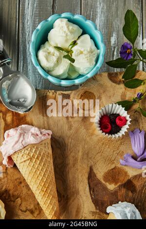 Gelato alla vaniglia, al mirtillo e al lampone in coni di waffle e gelato in tazza su tavola di legno con frutti di bosco e fiori. Foto Stock
