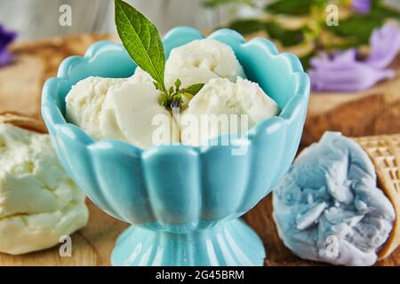 Gelato alla vaniglia, al mirtillo e al lampone in coni di waffle e gelato in tazza su tavola di legno con frutti di bosco e fiori. Foto Stock