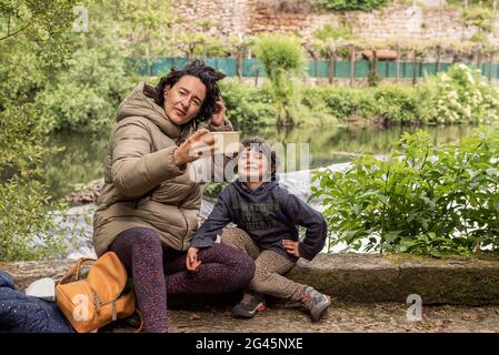 madre e sua figlia che prendono un selfie seduto accanto ad un fiume Foto Stock
