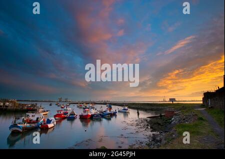 Piccole barche da pesca in un piccolo porto con i moli commerciali e un suggestivo cielo al tramonto sullo sfondo. Foto Stock