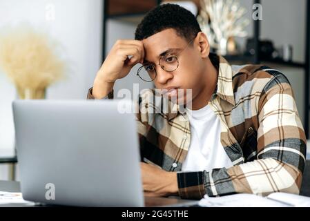 Sconvolto frustrato ragazzo ispanico, lavoratore d'ufficio o freelance, si siede alla scrivania, guarda tristemente il suo laptop, ha fallito l'accordo, ha avuto un cattivo risultato di lavoro, ha problemi, umore negativo, preoccupato Foto Stock