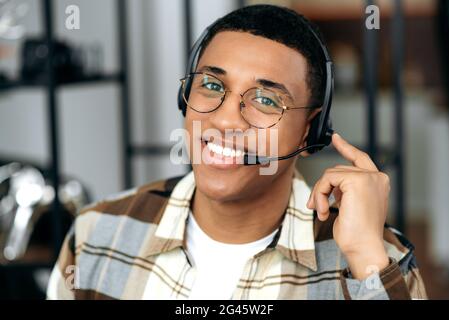 Primo piano di un bel giovane ispanico che indossa occhiali e cuffie, esperto, consulente o operatore, guarda direttamente alla fotocamera, sorridendo amichevole. Concetto di servizio di supporto, hot line Foto Stock