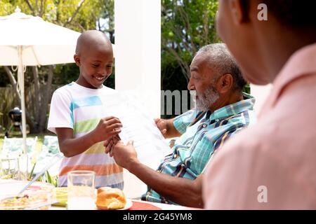 Ragazzo afroamericano che mostra al nonno i suoi compiti durante un pranzo di famiglia in giardino Foto Stock