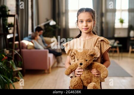 Ragazza carina con orsetto in piedi in soggiorno contro la sua famiglia Foto Stock