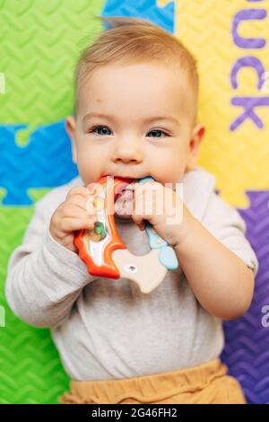 Il bambino piccolo in un bodysuit bianco tiene e gnaws un sonaglino luminoso sdraiato su un tappeto colorato Foto Stock