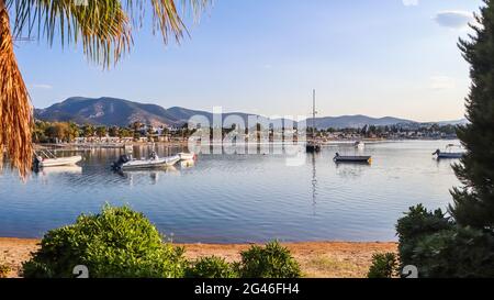 Bella Baia con acqua calma spiaggia di sabbia, barche e yacht. Piccola città e montagne verdi sulla costa del Mar Egeo Foto Stock