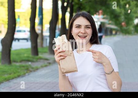 Una bella bruna ragazza mantiene uno shawarma in strada. Cibo sulla strada per lavorare. Pubblicità fast food cafe. Foto Stock