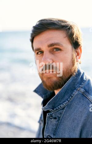 Ritratto di un uomo penoso semitirato in una giacca in denim sullo sfondo del mare. Primo piano Foto Stock