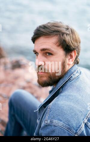 Uomo pensivo in jeans e una giacca in denim guarda in lontananza con la testa girata sullo sfondo del mare. Primo piano Foto Stock