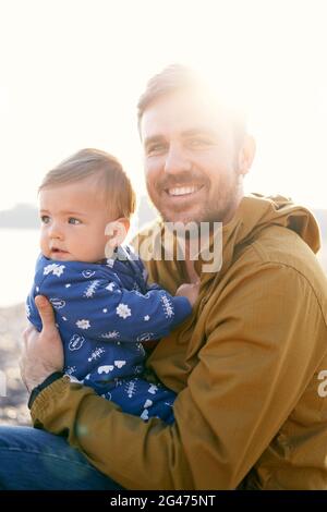 Il papà sorridente in una giacca marrone si siede e tiene un bambino in una tuta blu tra le braccia, girata lateralmente in piena luce del sole. C Foto Stock