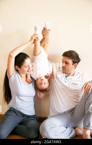 Mamma e papà stanno sollevando una bambina dalle gambe mentre si siedono sul pavimento Foto Stock