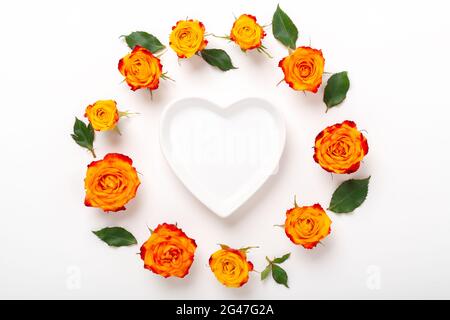 Composizione dei fiori. Corona fatta di fiori di rosa e ciotola vuota a forma di cuore su sfondo bianco. Concetto di giorno di San Valentino. Disposizione piatta, vista dall'alto - immagine Foto Stock