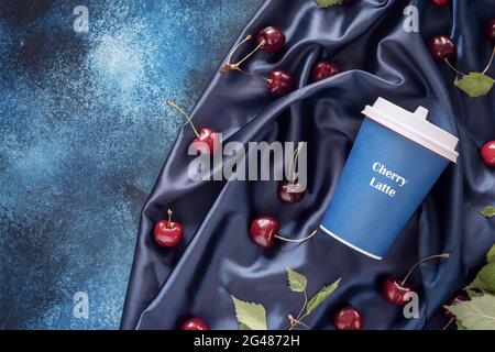 Composizione alimentare con tazza artigianale di caffè, ciliegie e foglie su sfondo blu, satinato, piegato con spazio di copia. Latte di ciliegia, nuovo tipo di caffè Foto Stock