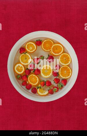 sfondo rosso testurizzato con una grande ciotola di arance e fragole che galleggiano in acqua per un bagno di frutta Foto Stock