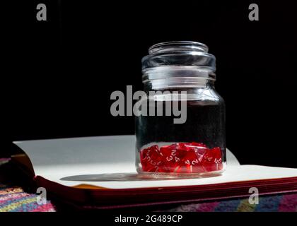 Un vaso di vetro riempito con dadi rossi su 4 lati su un notebook aperto alla luce del sole Foto Stock