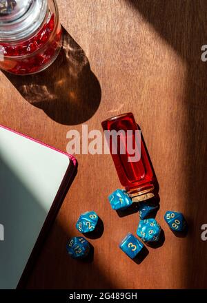 Immagine verticale di una serie di dadi da gioco di ruolo in marmo blu, una bottiglia di vetro con liquido rosso, un taccuino e un vaso di vetro riempito di dadi rossi Foto Stock