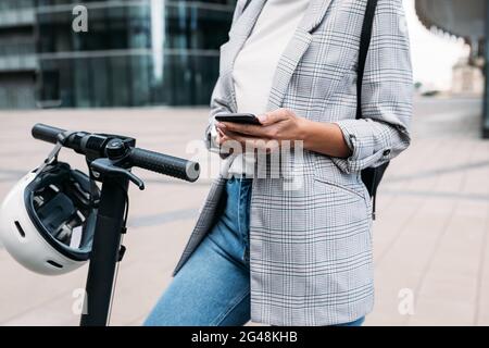 Scatto corto di femmina in casuals usando un'applicazione su un telefono cellulare per sbloccare lo scooter elettrico a spinta Foto Stock