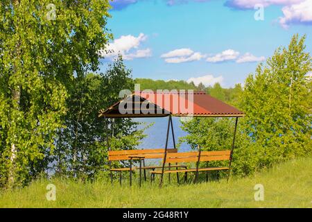 Paesaggio estivo con pergolato in legno antico, gazebo. Panchine di legno sotto un tetto di tegole rosse su una riva di un fiume, lago. Erba verde e alberi contro il blu Foto Stock