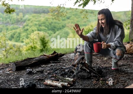 Una giovane donna si riscalda vicino al fuoco con una tazza di bevanda calda nella foresta. Foto Stock