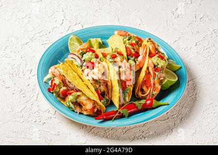 conchiglie di taco con gamberi alla griglia, insalata di cavolo, lime e guacamole su un piatto blu su un tavolo bianco Foto Stock