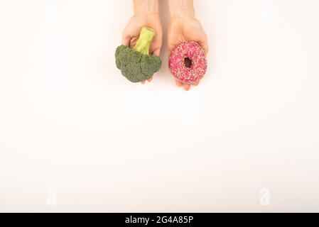 Confronto delle abitudini alimentari. Donna che tiene broccoli e ciambella su sfondo bianco. Spazio di copia Foto Stock
