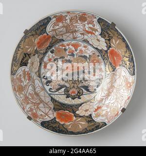 Piatto con vaso Flower, piante fiorite e rotoli floreali. Piatto di porcellana, dipinto in blu e sullo smalto rosso e oro. Sul piatto un vaso di fiori con crisantemi e peonie; sul vaso un loto stilizzato. La parete e il bordo sono divisi in sei compartimenti con varie piante in fiore dietro una recinzione e riflessi di peonia su uno sfondo blu. La parete esterna con tre rami di fiori. (Crisantemo, Prunus, Peonia). La parte inferiore con preen. Il piatto viene impigliato in un telaio di stampo. Imari. Foto Stock
