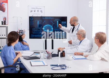 Medico che indica la radiografia medica ai colleghi dell'ospedale analizzando la presentazione della malattia cerebrale utilizzando l'alta tecnologia nella sala riunioni. Il team ospedaliero monitora il trattamento malato per le competenze sanitarie Foto Stock