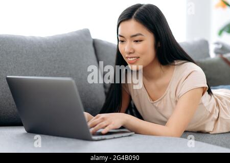Una ragazza asiatica carina e positiva si trova a casa sul divano dello stomaco, utilizza un computer portatile, naviga in Internet, chattando sui social network, guarda un webinar online, scrivendo su e-mail, sorridendo Foto Stock