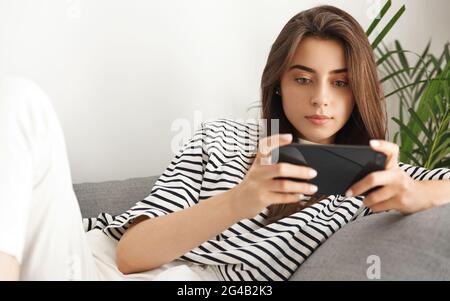 Giovane donna attraente sdraiata sul divano e guardando i video sullo smartphone. Ragazza che tiene il telefono cellulare orizzontale, guardando lo schermo, giocando un videogioco Foto Stock