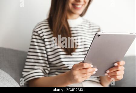 Ripresa di una donna sorridente che usa un tablet, una ragazza che legge o guarda online smth sul suo gadget mentre si siede a casa sul divano Foto Stock