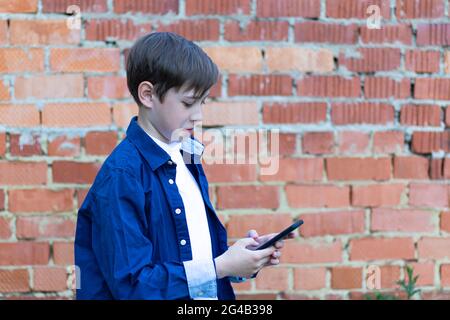 Un ragazzo adolescente in una camicia blu con uno smartphone in mano comunica con gli amici sui social network sullo sfondo di un muro di mattoni. Sele Foto Stock