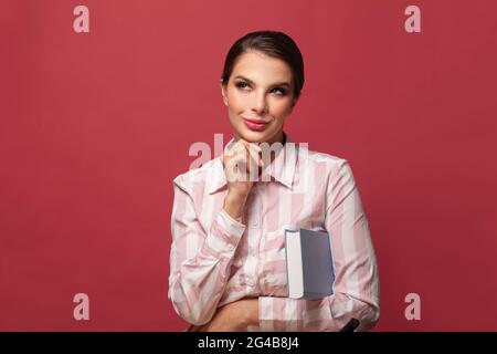 Sorridente giovane bruna donna che tiene il libro in posa isolato su sfondo rosso muro, studio ritratto. Spazio di copia fittizio. La gente sincera emozioni di vita Foto Stock