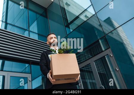 Infornato lavoratore di ufficio maschile depresso con scatola di personale roba che cammina all'aperto, ha perso lavoro e lasciato senza soldi Foto Stock