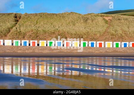 Capanne bianche, con porte colorate, si trovano di fronte alle dune con riflessi nella sabbia bagnata della spiaggia di Woolacombe Beach, Devon Foto Stock