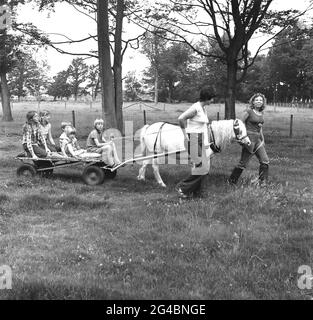 Anni '80, storico, vacanza divertimento per tutta la famiglia in una fattoria, alcuni bambini piccoli che hanno un giro su un piccolo trolley piano a quattro ruote o piattaforma, trainato da un cavallo intorno a un campo, guidato da una lavoratrice agricola femminile e una madre accanto, Hortle, Hampshire, Inghilterra, Regno Unito. Foto Stock