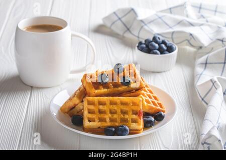 Waffle belgi con mirtilli e caffè su un tavolo di legno. Bevanda calda del mattino e frutti di bosco, concetto di colazione. Latte in una tazza bianca e primi piatti di pasta Foto Stock