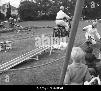 anni '60, storico, fuori in un campo, un membro di un team di esposizione moto che esegue una stunt di saltare sopra le lattine di olio sulla sua moto. Le mostre acrobatiche di motociclette ai carnevali esterni e gli eventi speciali sono stati popolari in quest'epoca, con i Royal Signals, i "White Helmets", un team di esposizione dell'esercito britannico, i più noti. Avevano eseguito la prima mostra registrata di stunt nel 1928, mentre i piloti di spedizione militari, che erano una parte chiave delle comunicazioni dell'esercito in questo momento, avevano bisogno di essere motociclisti altamente qualificati. Foto Stock