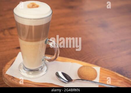 Caffè latte macchiato in una tazza di caffè trasparente. La tazza è su uno sfondo di legno con il biscotto di caffè sul tavolo accanto al c Foto Stock
