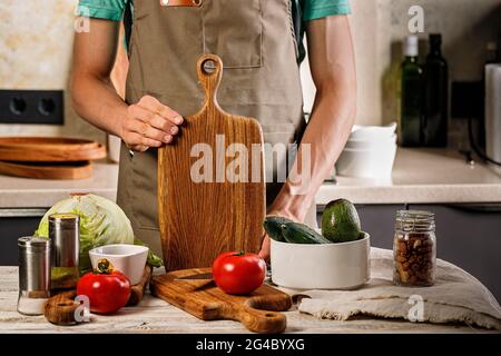 Un giovane chef in un grembiule marrone tiene un tagliere di legno. Le verdure per un'insalata vegetariana sono sul tavolo. Cucina cibo delizioso e sano in Foto Stock