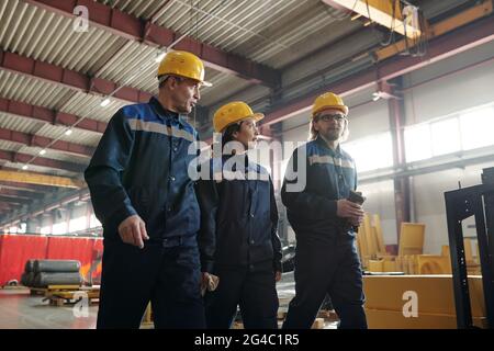 Lavoratori di fabbrica sicuri in giallo hardhats camminare insieme in officina industriale e discutere i processi di lavoro Foto Stock